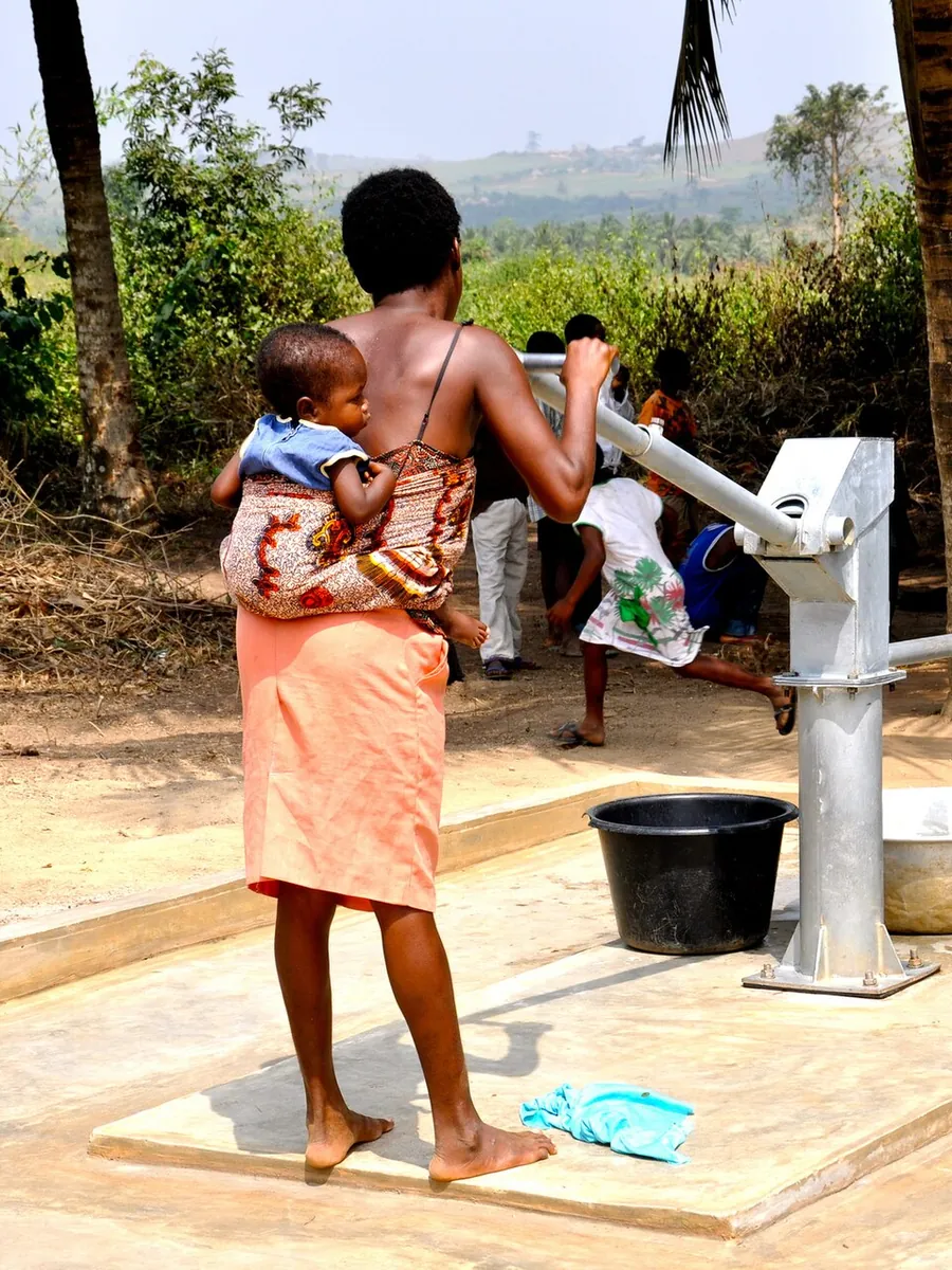 A mother carrying her baby fetches clean water at a village pump while children fill basins