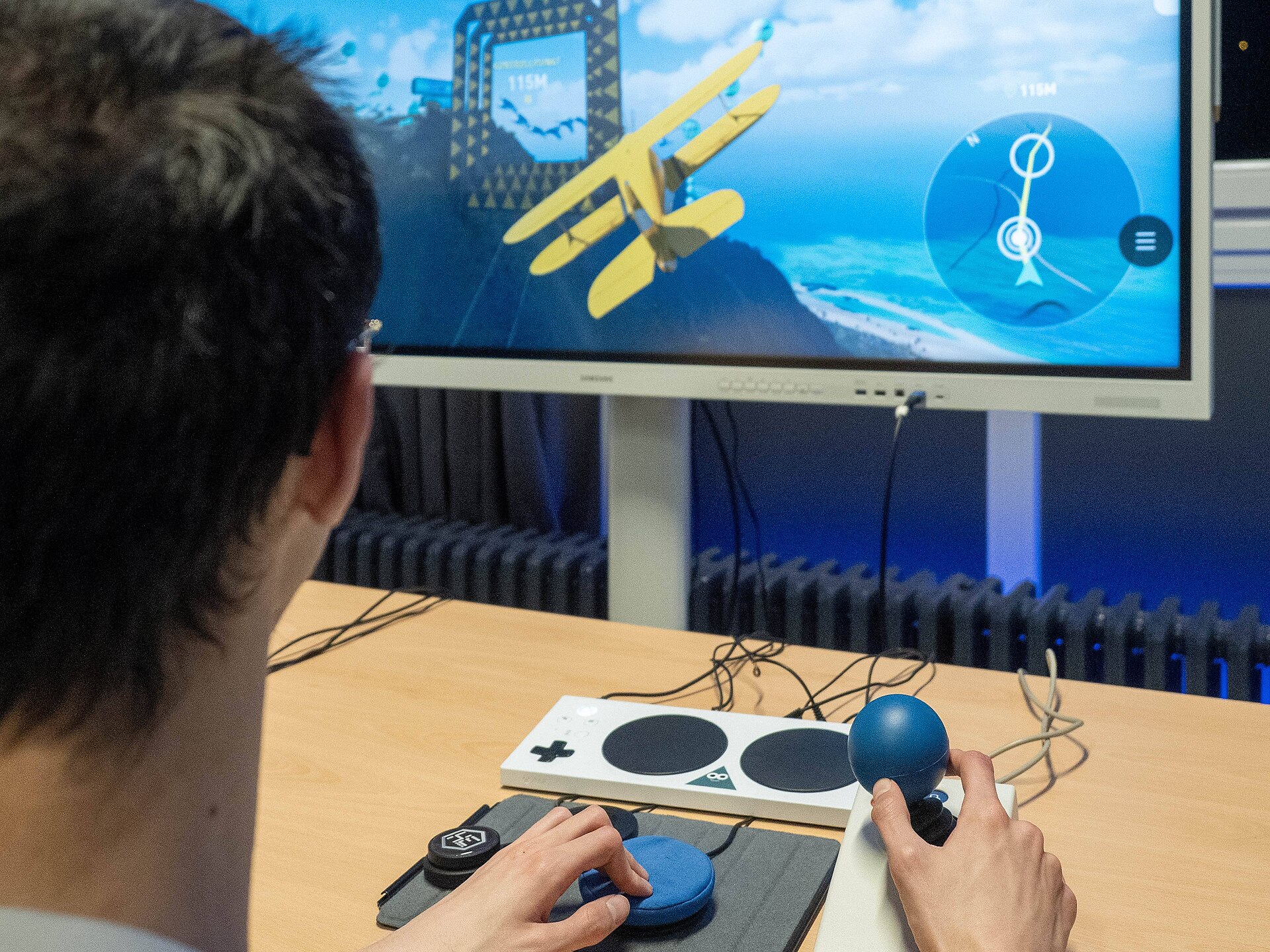A person uses an adaptive game controller and a large joystick to fly a yellow biplane in a game on a large screen